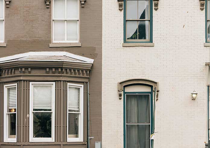 The front of two houses are shown, the left shows a brown painted front with an angled bay window, the right house has been painted cream with green window frames.