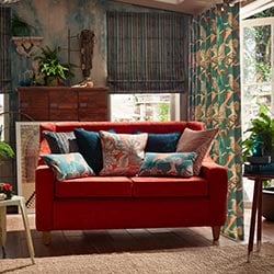 A green velvet striped roman blind sits in a conservatory room. On the right side a green curtain with cranes is on the french doors. A red velvet couch is centre of the room.