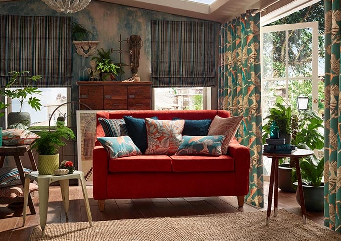 A green velvet striped roman blind sits in a conservatory room. On the right side a green curtain with cranes is on the french doors. A red velvet couch is centre of the room.