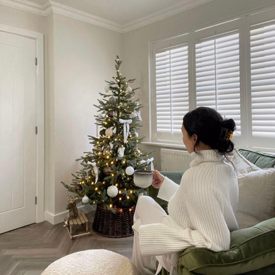 Woman sat infront of a Christmas tree in a living room with white wooden shutters.