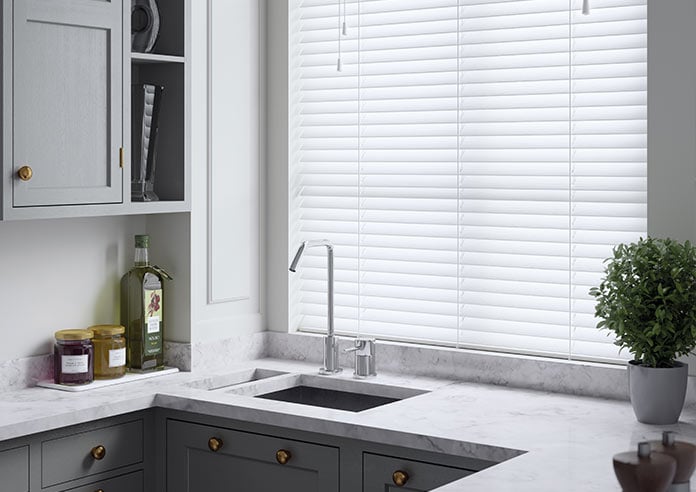 A white faux wooden blind adorns a window in a kitchen.  A small plant is on the right of a marbled, grey-toned sink area.