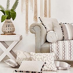 Cosy soft neutral cushions, on the floor next to a grey sofa.  A cream vase with green plants sit on a table next to it.