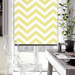 A zigzag, striped yellow roller blind is shown in an modern home office.  The desk is grey with a plant on it, and a clear seat is in the bnottom left hand corner.