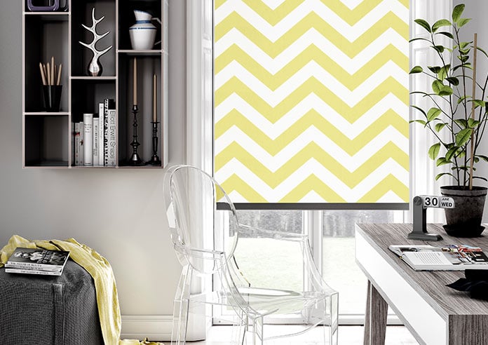 A zigzag, striped yellow roller blind is shown in an modern home office.  The desk is grey with a plant on it, and a clear seat is in the bnottom left hand corner.