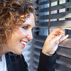 An image of amy wilson looking through her grey taped wooden blinds