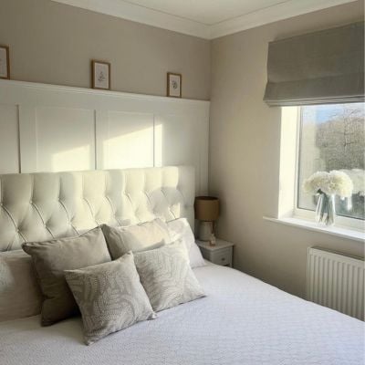 A neutral bedroom featuring a white panelled wall and upholstered bed, with greige, thick Roman blinds placed outside the window recess. 