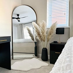 An Image showing a bedroom with a wooden blind in the window, grey drawers and a large mirror