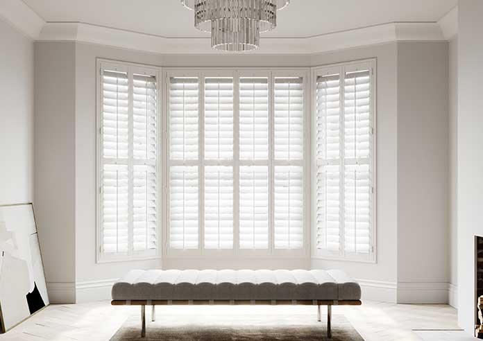 White Shutters are shown in a living room bay window. The room is neutral using white and grey tones.