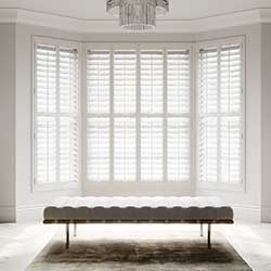 White Shutters are shown in a living room bay window. The room is neutral using white and grey tones.