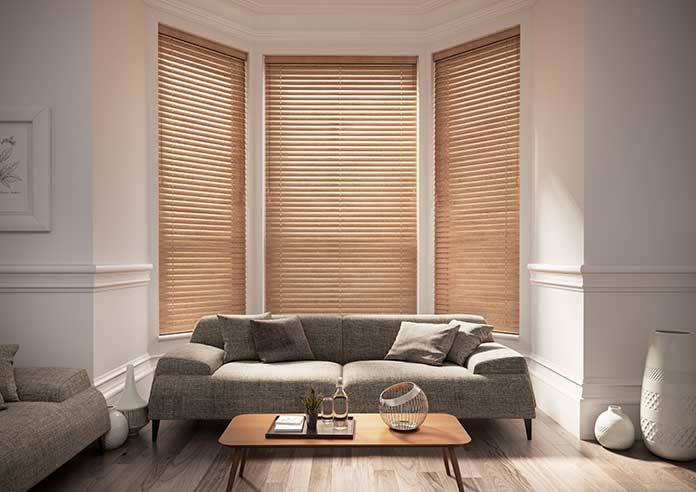 Real wooden blinds are shown in a living room bay window. The walls are cream, the sofa is grey and the room sparsely decorated.