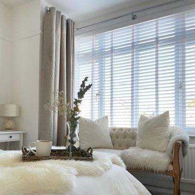 White, taped, wooden Venetian blinds paired with linen-look beige curtains in a bedroom 